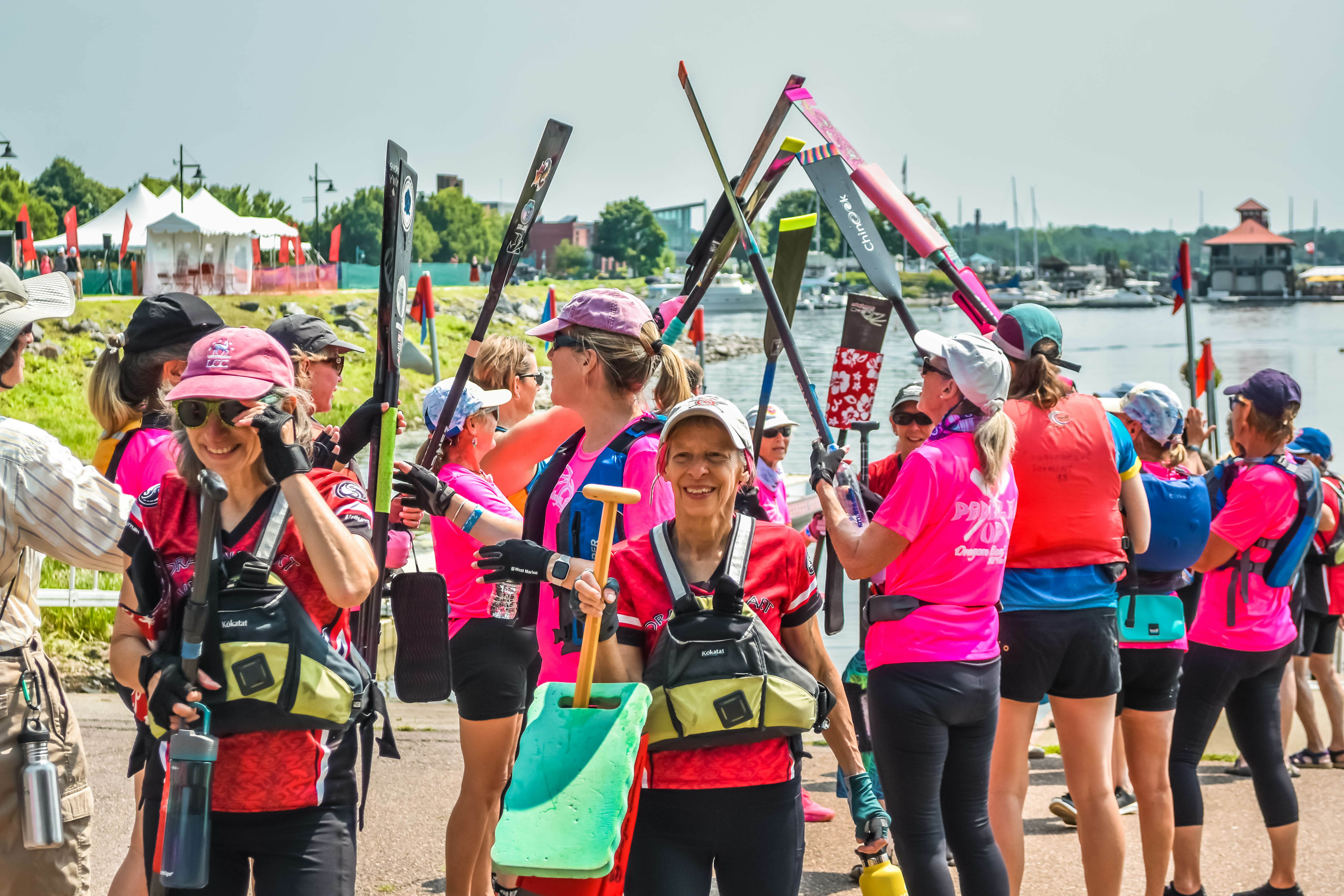 A diverse group of women in pink shirts, holding paddles near a waterfront.