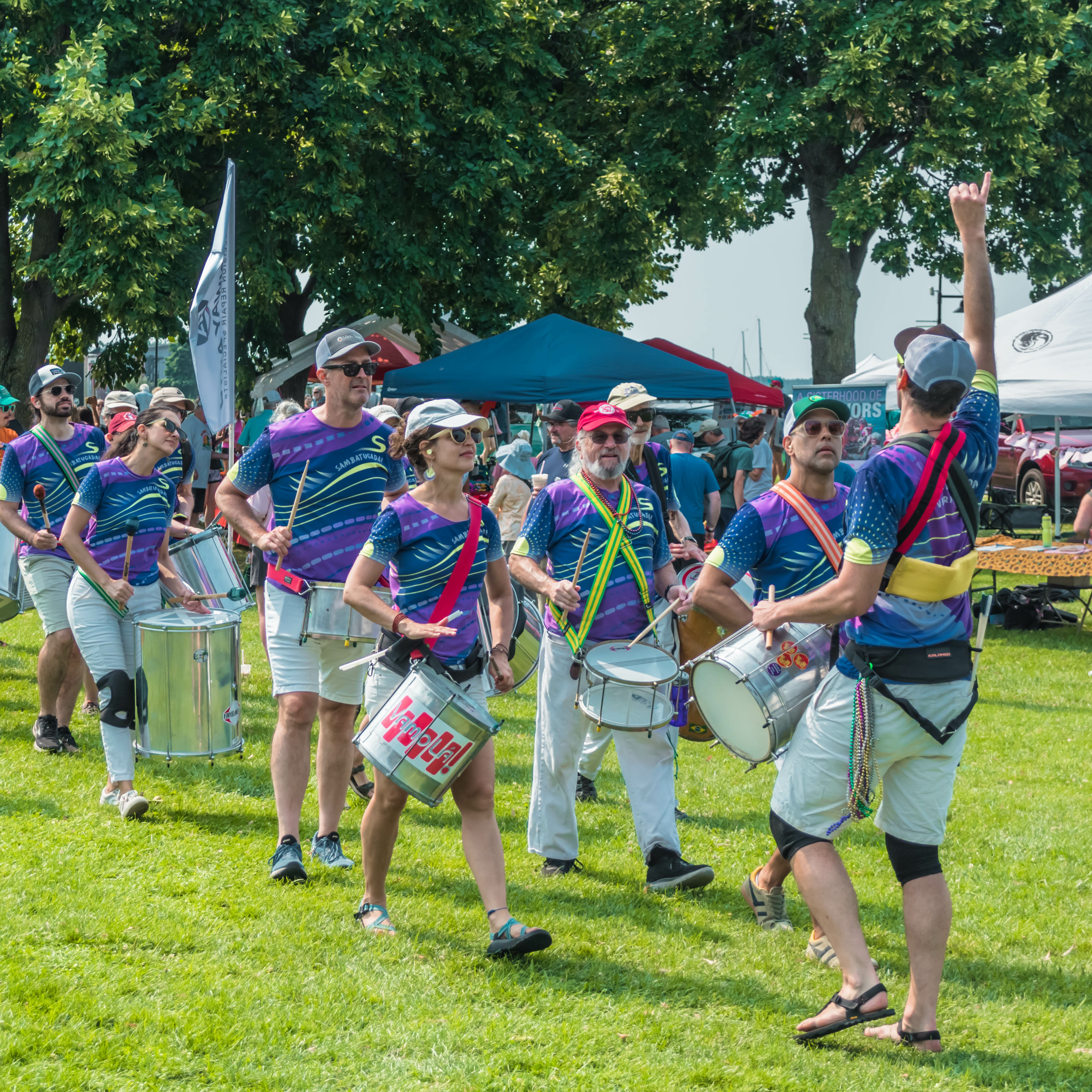 A drumline in colorful outfits performs outdoors, surrounded by tents and spectators.