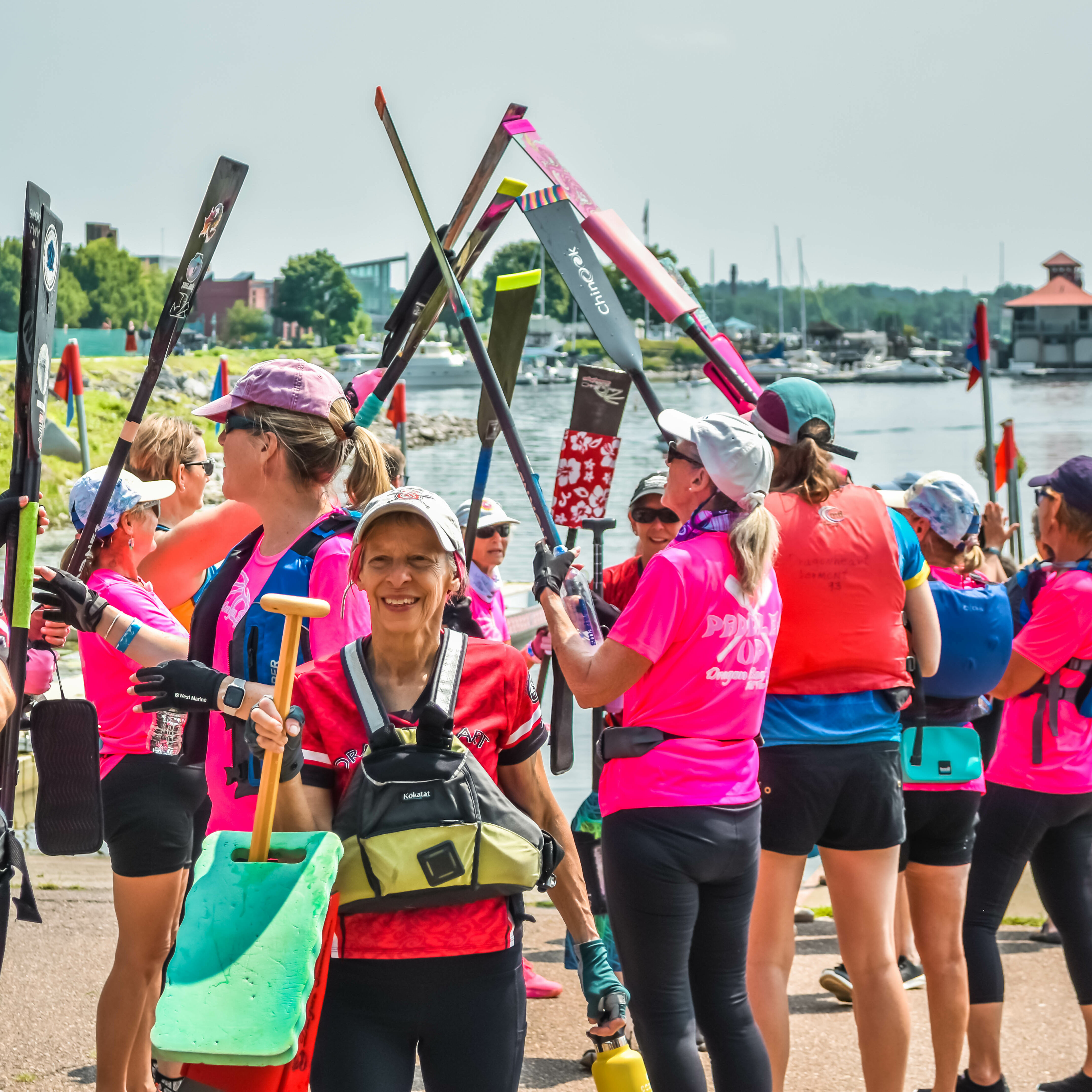 Women in bright shirts hold paddles and celebrate by the water's edge.