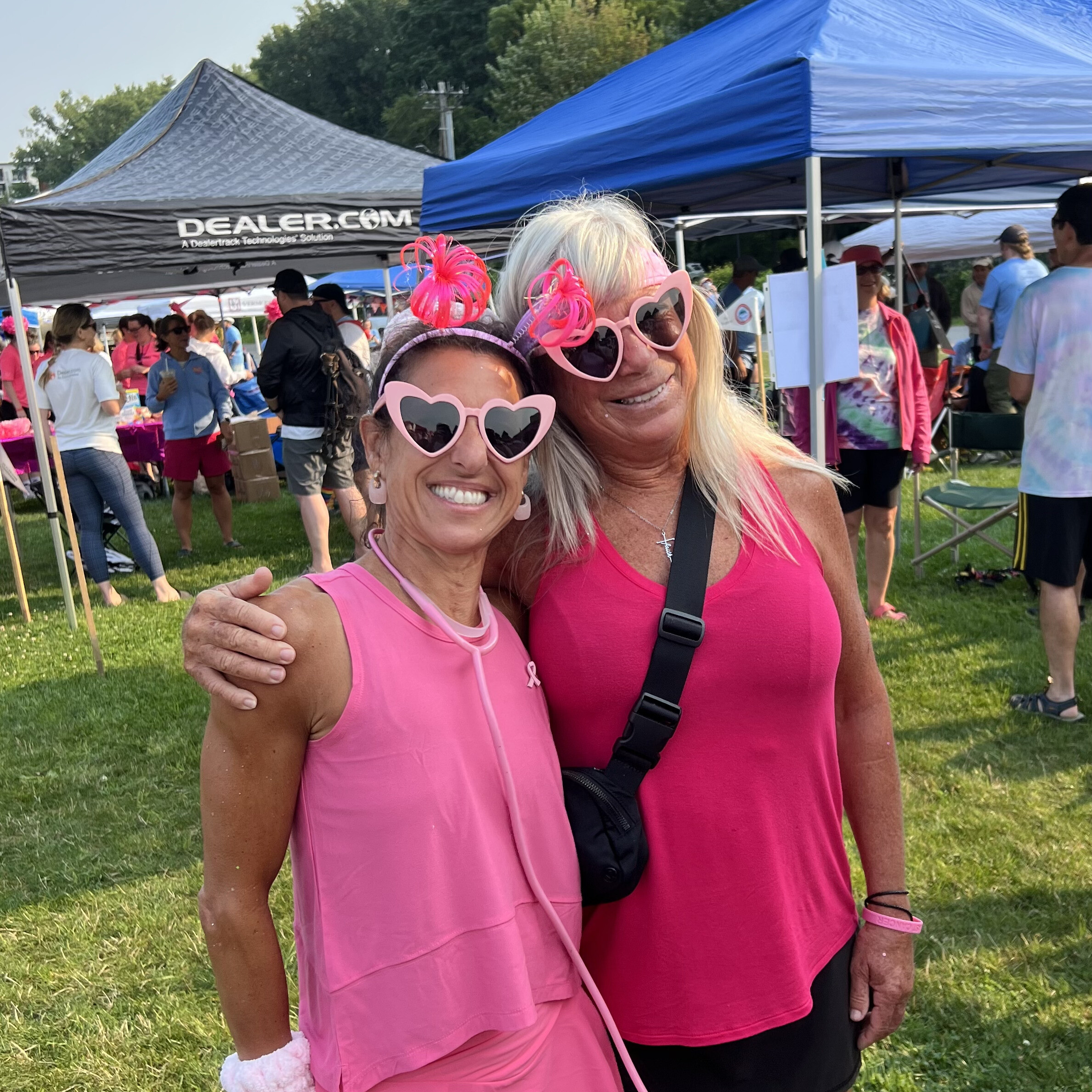 Two women in pink attire and heart-shaped sunglasses smile together at an outdoor event.