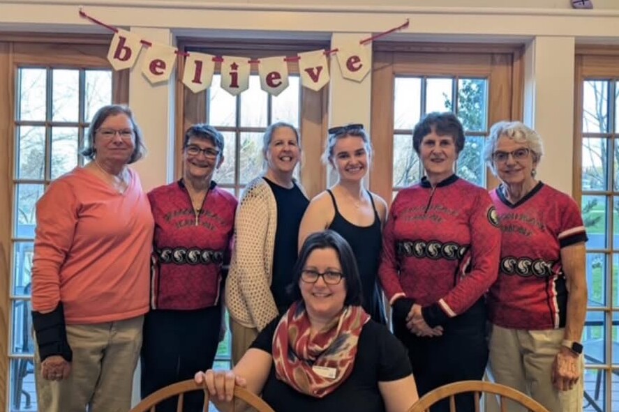 Seven women posed together inside a bright room, smiling, with a "believe" banner overhead.