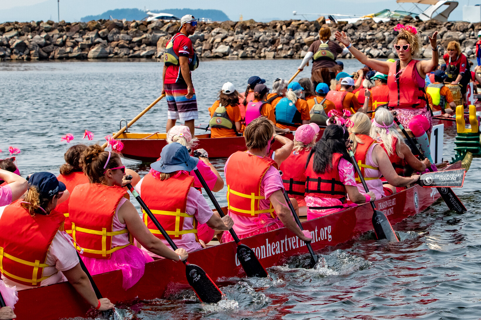 Dragon boat participants in colorful life vests paddling on water, led by a cheerleader.