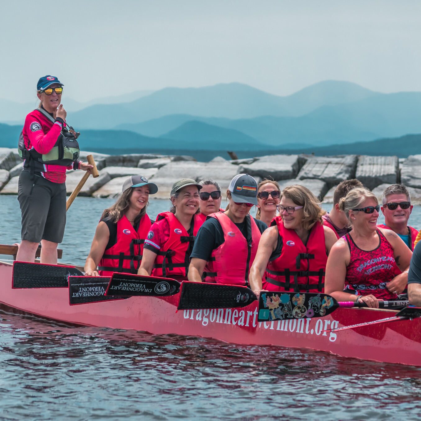 A dragon boat crew in pink life jackets smiles while paddling on a lake with mountains in the background.