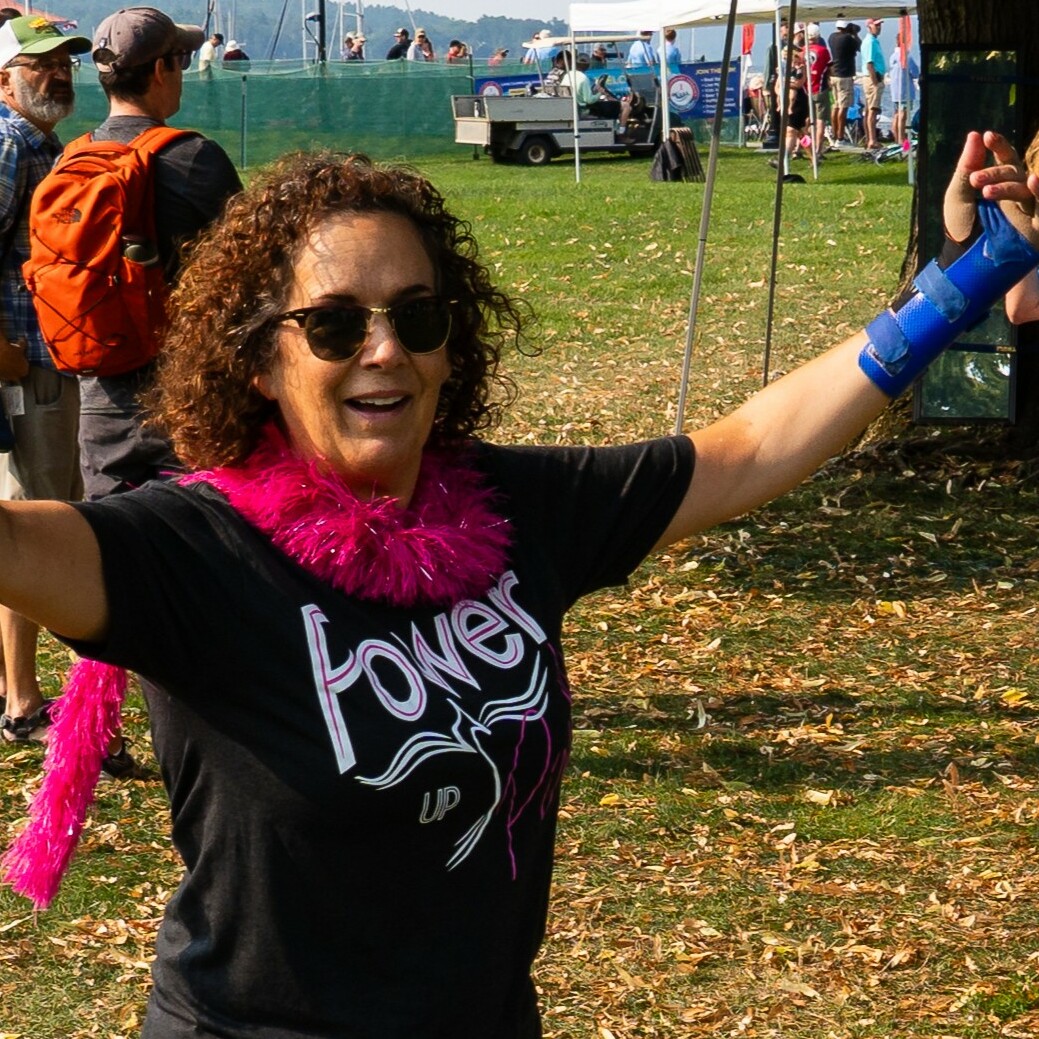 Woman in a black "Power Up" shirt with pink boa, dancing at an outdoor event.