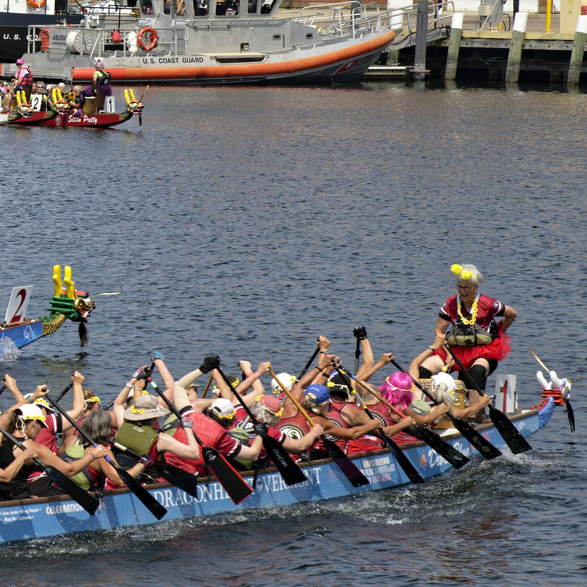 Teams compete in a dragon boat race on a sunny day by a docked Coast Guard boat.